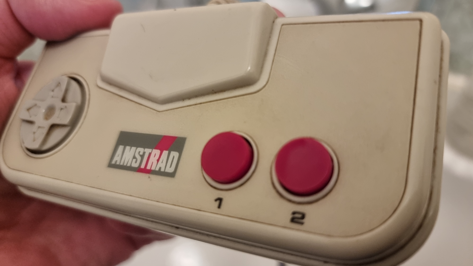 The image is a close-up of an Amstrad game controller, showing signs of wear on its beige surface. It features a directional pad on the left, two red action buttons labeled "1" and "2" on the right, and the Amstrad logo across the center.