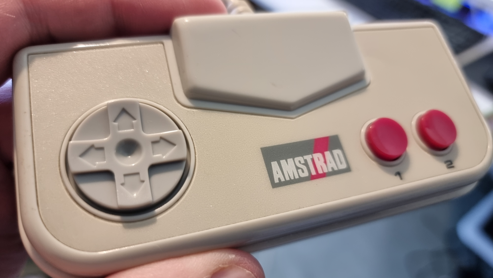 The image displays a close-up of a hand holding a restored and cleaned Amstrad GX4000 game controller. The controller looks in excellent condition with its directional pad, two red buttons, and the Amstrad logo prominently displayed. The restoration appears to have returned the controller to a state close to its original appearance.