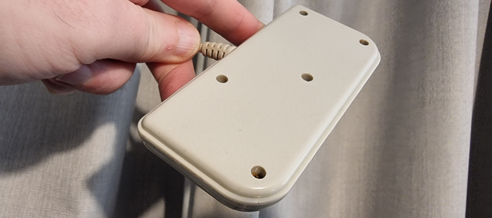 The image shows a hand holding the underside of a restored and cleaned Amstrad GX4000 game controller, displaying the screw holes for internal access and the clean, off-white finish of the controller's casing.